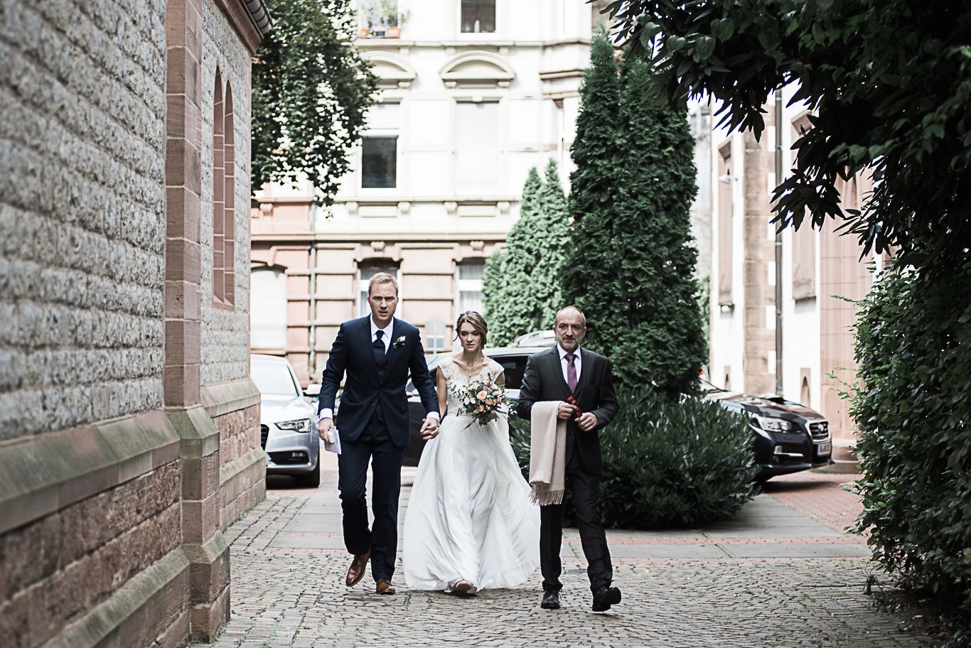 Brautpaar geht mit Vater durch eine Gasse zur Kirche