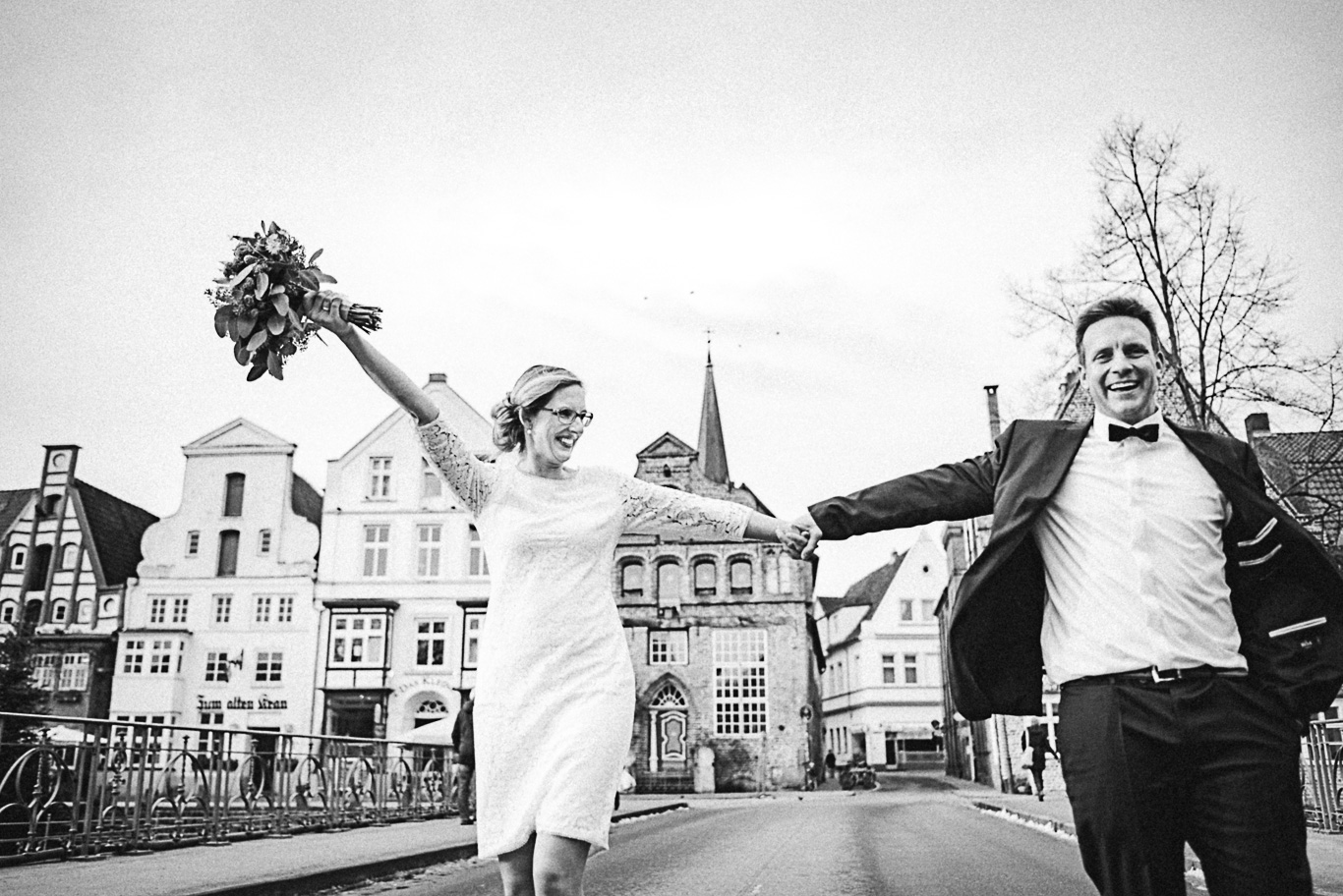 Braut streckt jubelnd den Arm mit Blumenstrauß vor Backsteinhäusern, Schwarzweißaufnahme