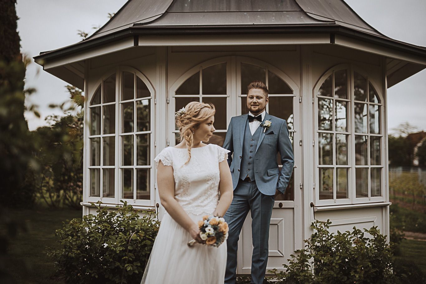 Brautpaar steht lächelnd vor Pavillon mit Blumenstrauß