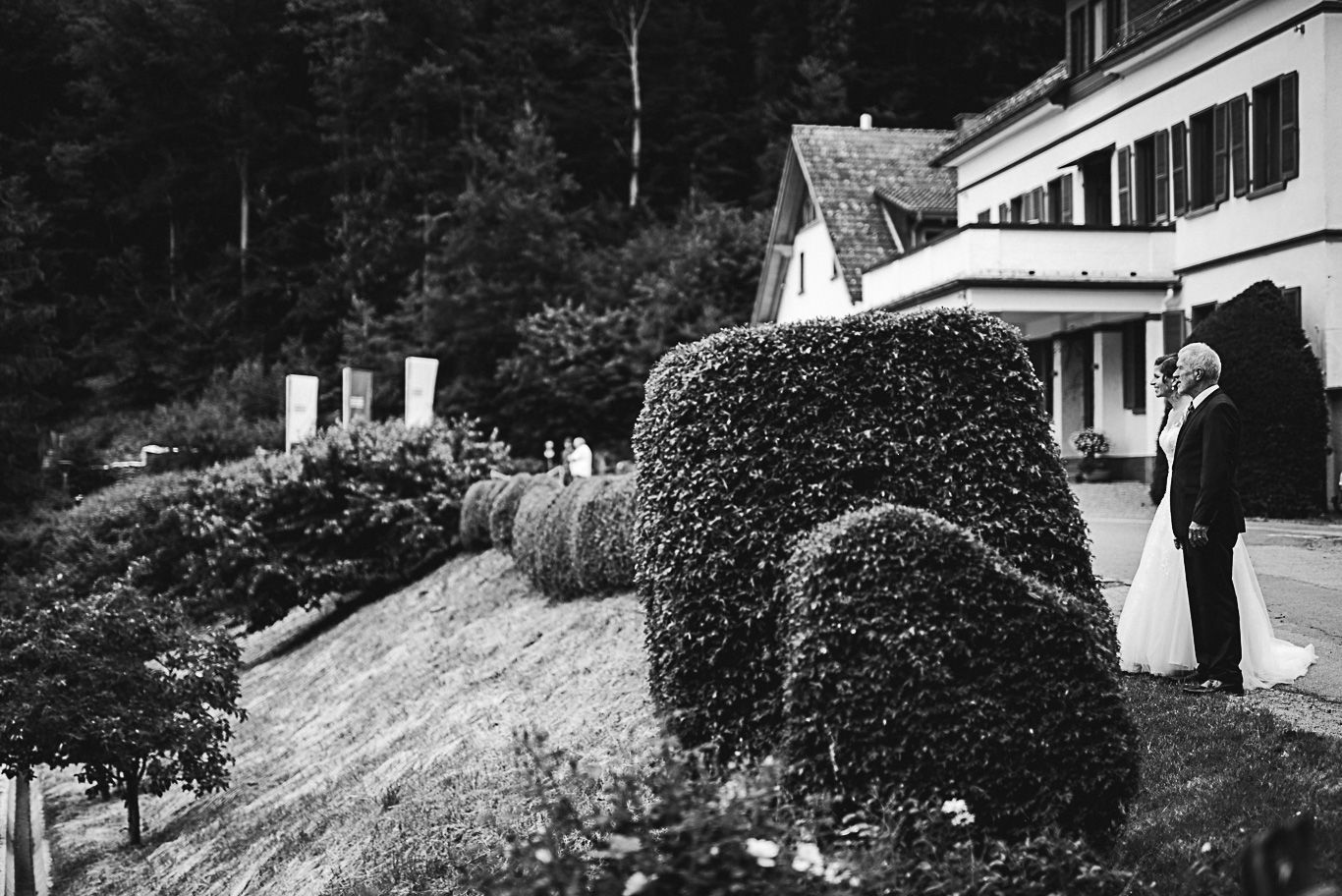 Brautpaar steht vor dem Hofgut zwischen Sträuchern, Schwarzweißaufnahme