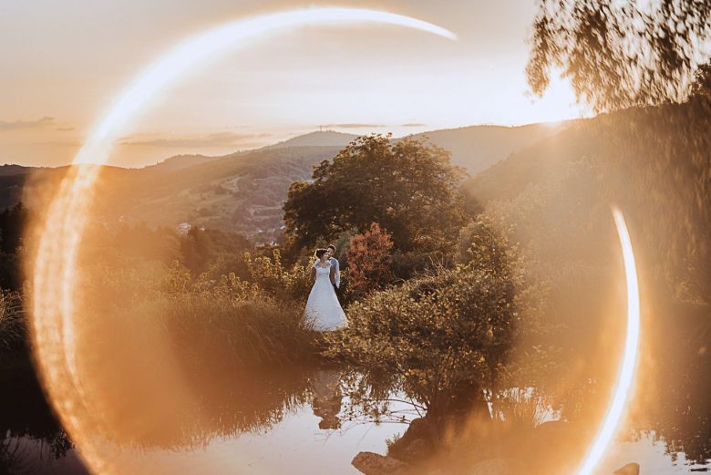 Braut steht klein zwischen Bäumen mit Lens-Flare-Effekt im Sonnenlicht