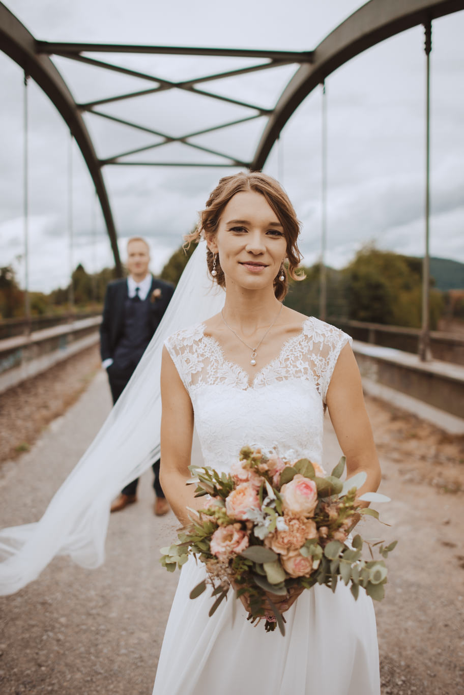Braut steht mit Blumenstrauß auf einer Brücke, Gäste im Hintergrund