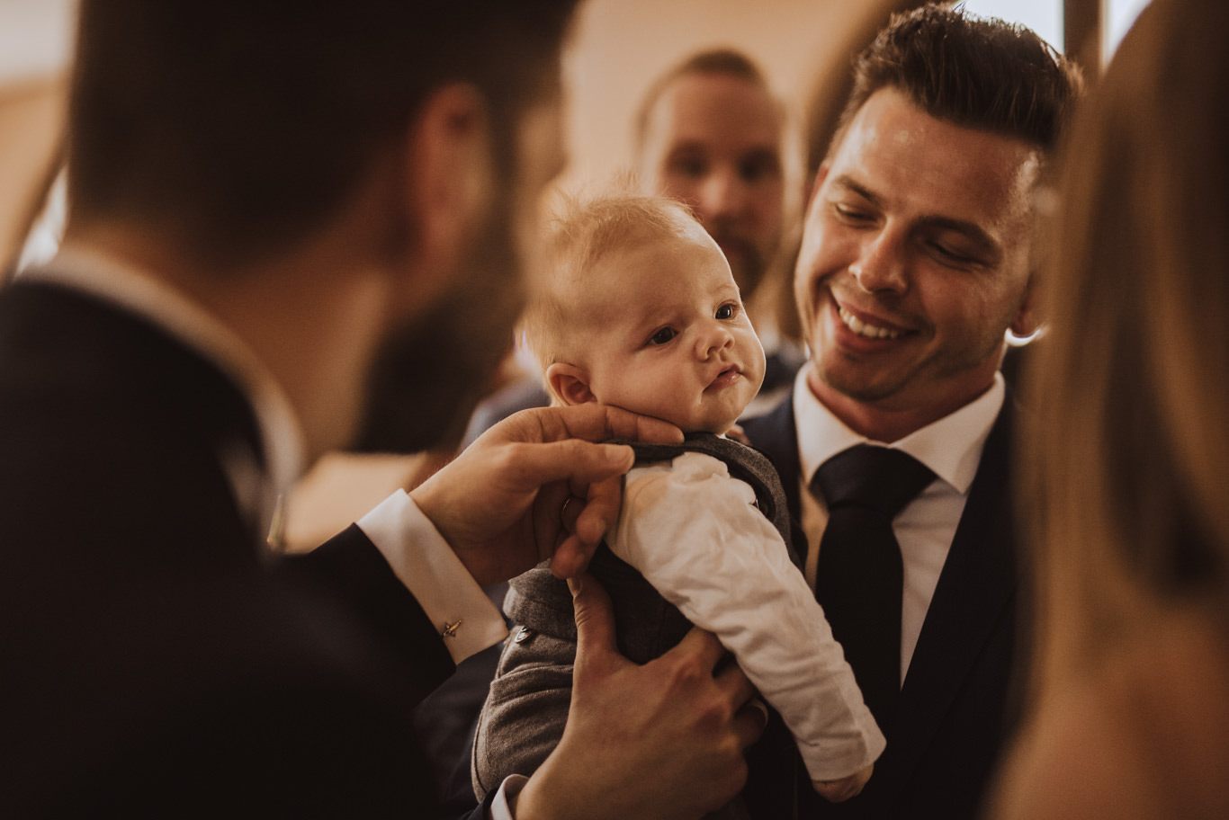Bräutigam hält lachend ein Baby vor Gästen im Arm