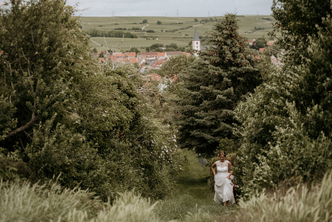 Braut läuft durch grüne Sträucher mit Blick auf Wachenheim
