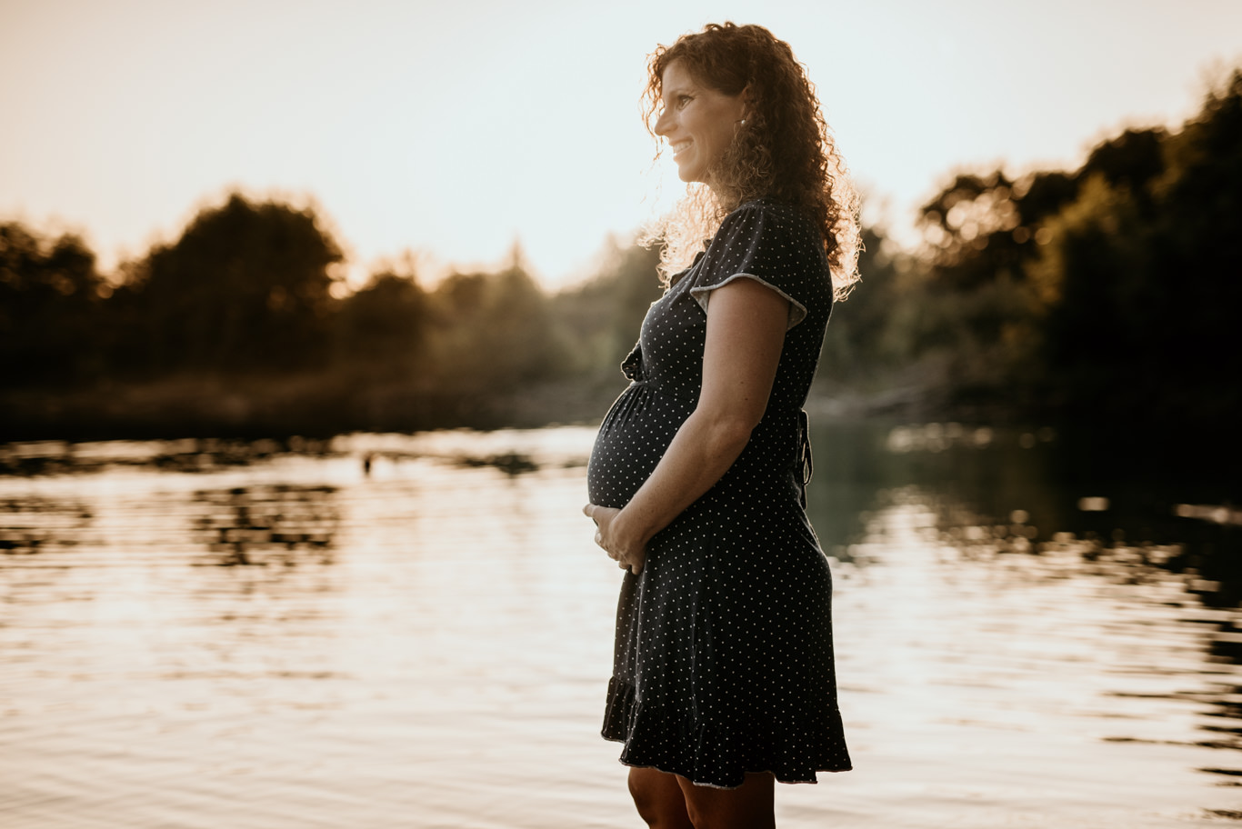 Schwangere steht im Gegenlicht am Ufer und hält ihren Bauch