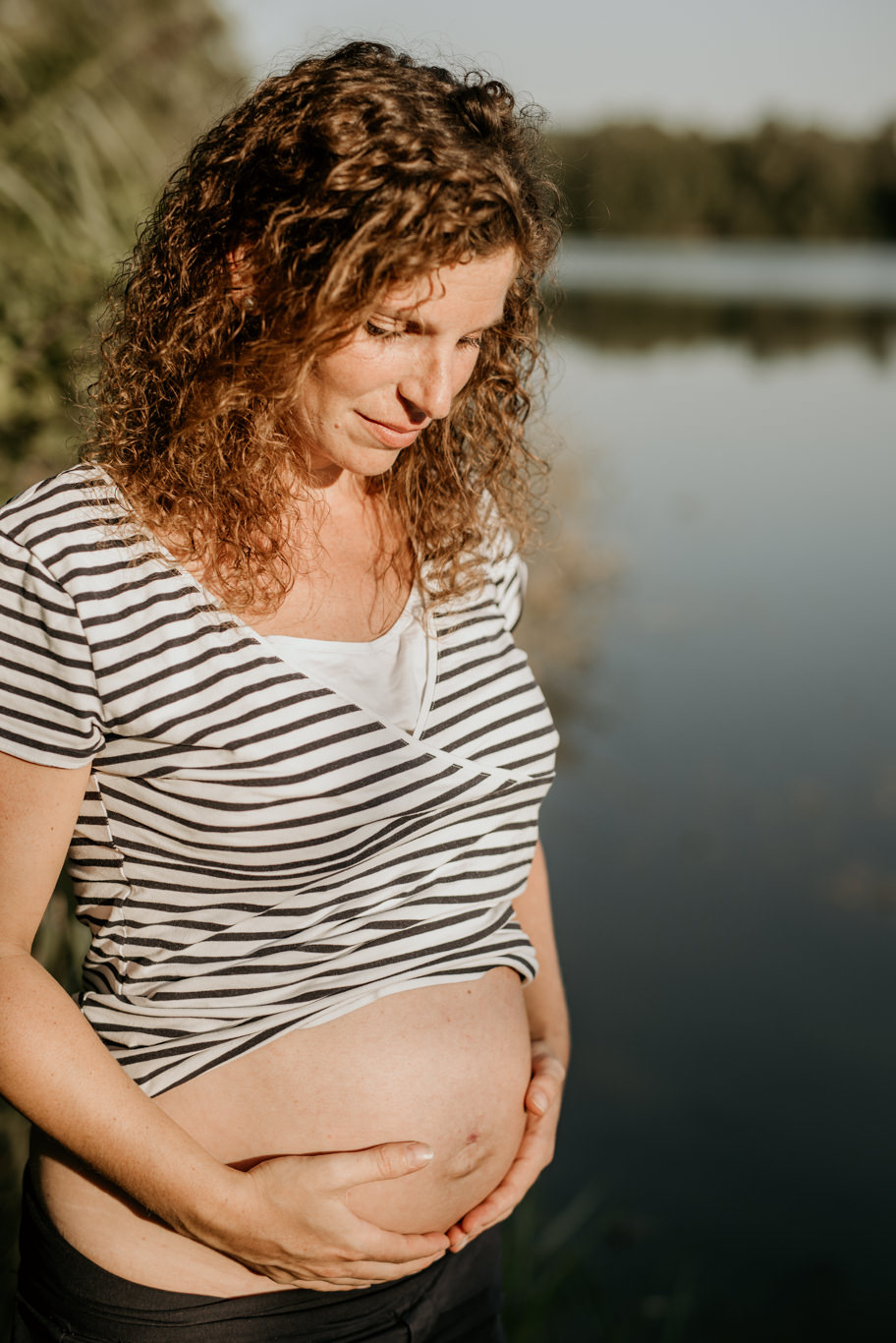 Schwangere hält ihren Bauch mit beiden Händen am Seeufer