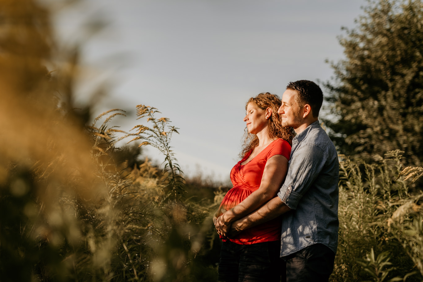 Schwangere und Partner stehen Hand in Hand im hohen Gras bei Sonnenuntergang