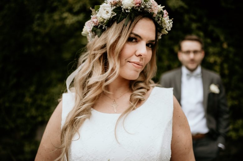 Braut mit Blumenkranz blickt lächelnd zur Seite, Bräutigam im Hintergrund