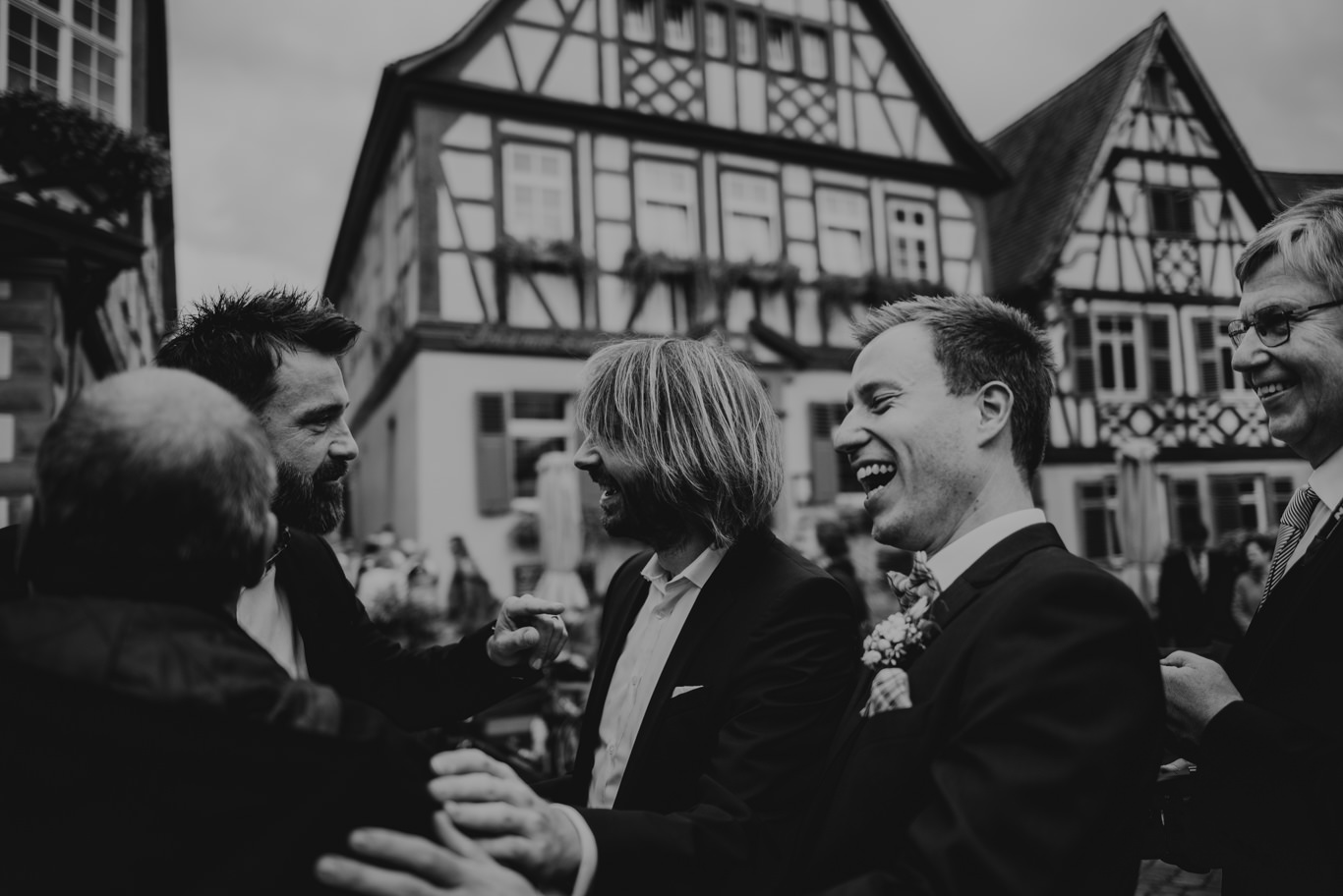 Bräutigam lacht mit Gästen vor Fachwerkhaus, Schwarzweißaufnahme