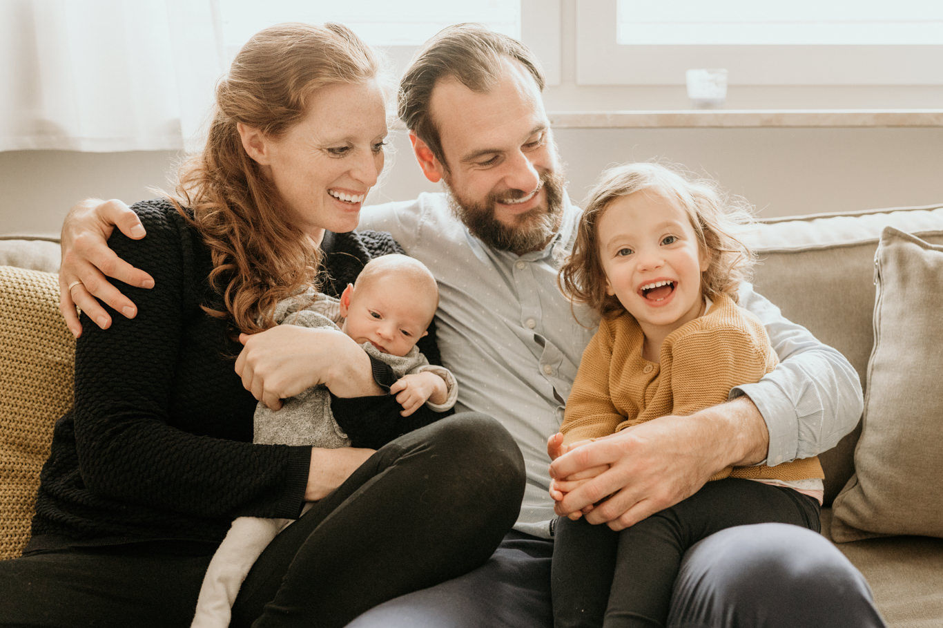 Familie lacht gemeinsam mit Neugeborenem und Tochter auf der Couch