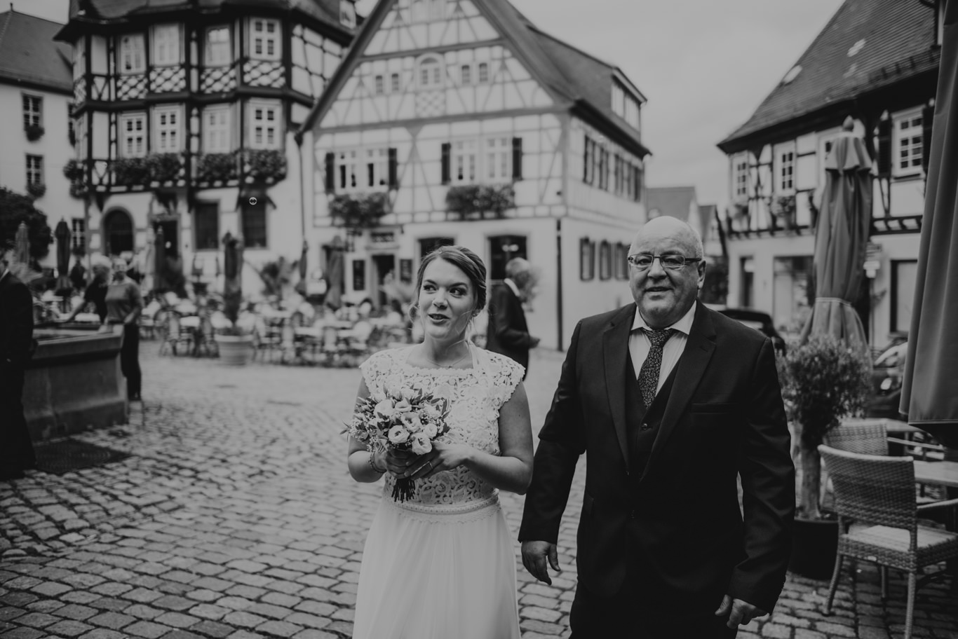 Brautvater führt Braut über Kopfsteinpflaster am Marktplatz, Schwarzweißaufnahme