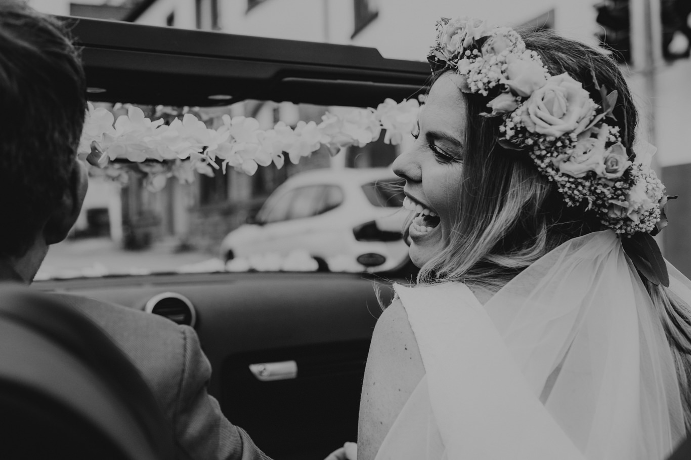 Braut mit Blumenkranz lacht im Fahrtwind des Cabrios