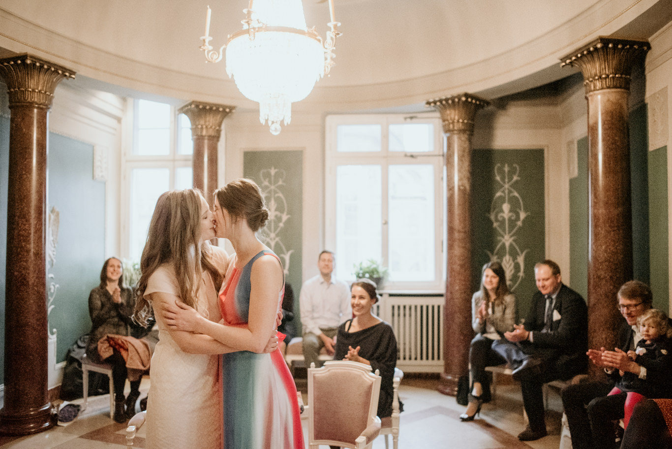 Zwei Bräute umarmen sich lächelnd vor Gästen im Trauzimmer