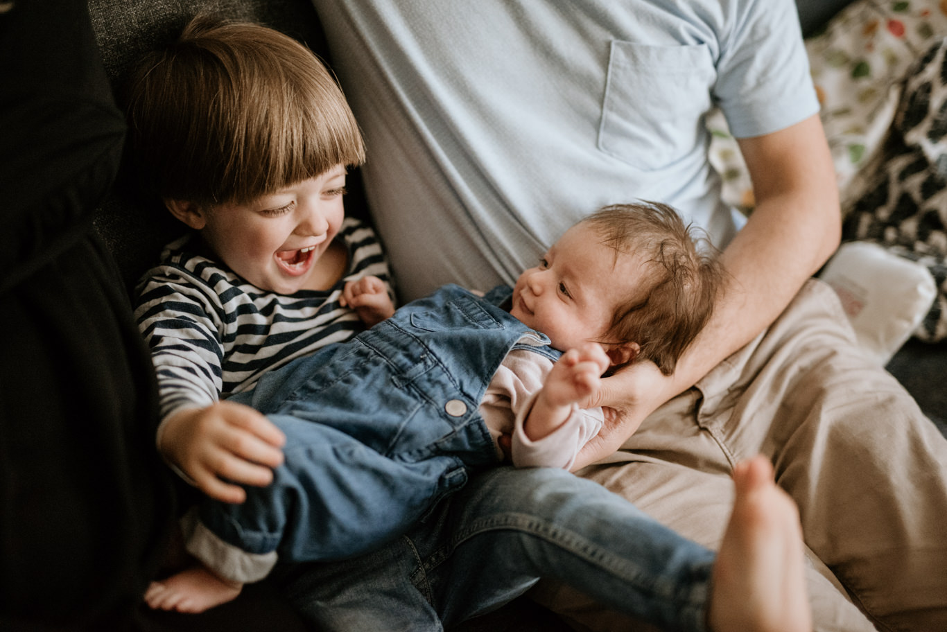 Vater hält das Baby, der große Bruder lacht ausgelassen daneben