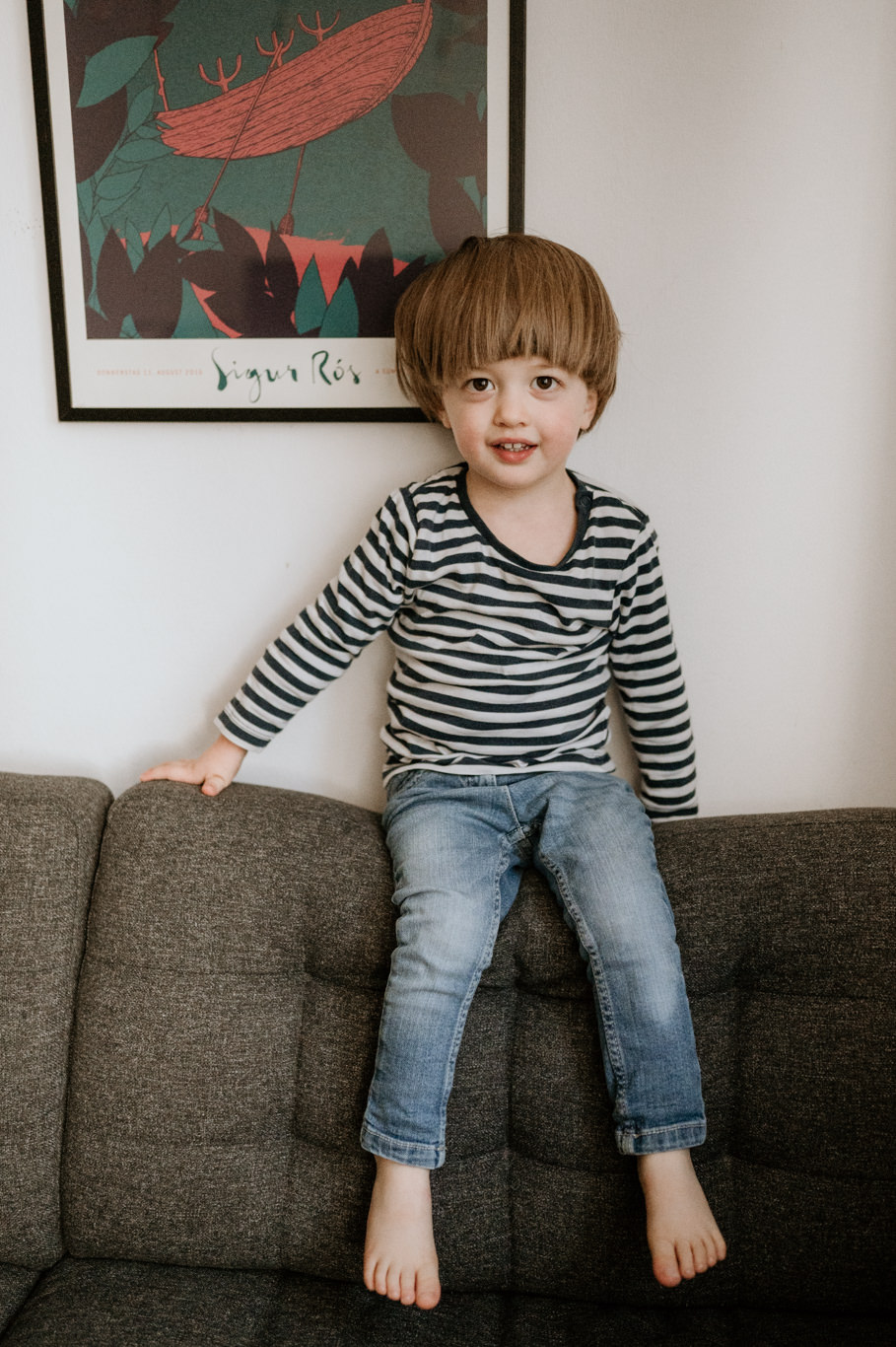 Junge im Streifenshirt sitzt barfuß lachend auf grauem Sofa
