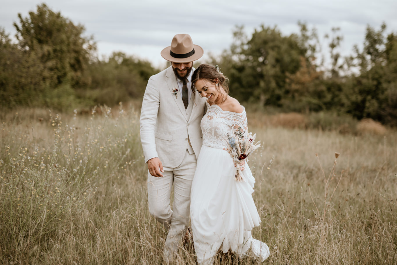 Boho-Brautpaar mit Hut geht Hand in Hand durch hohes Gras