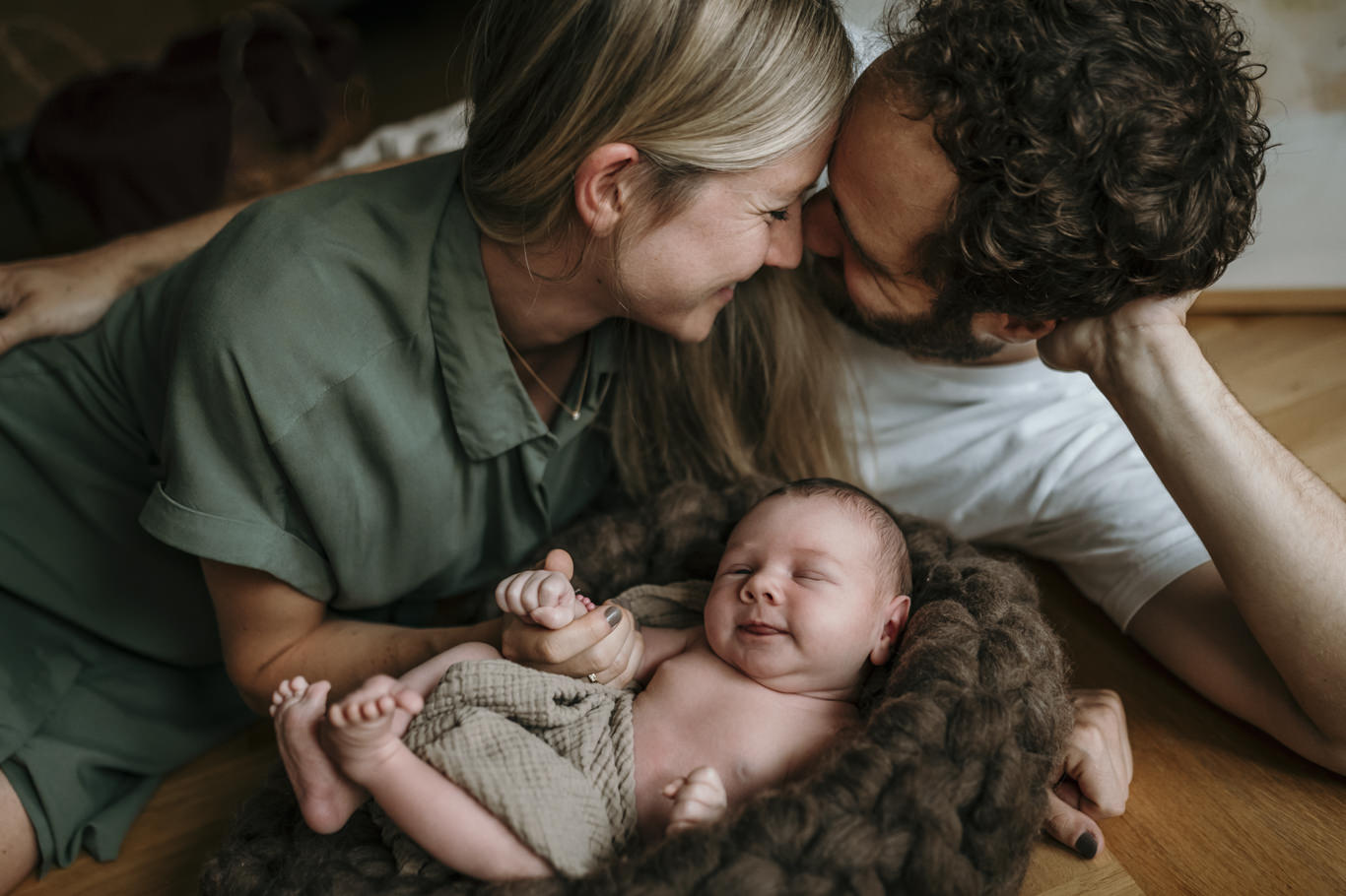 Eltern küssen sich lächelnd neben ihrem lachenden Baby auf dem Boden