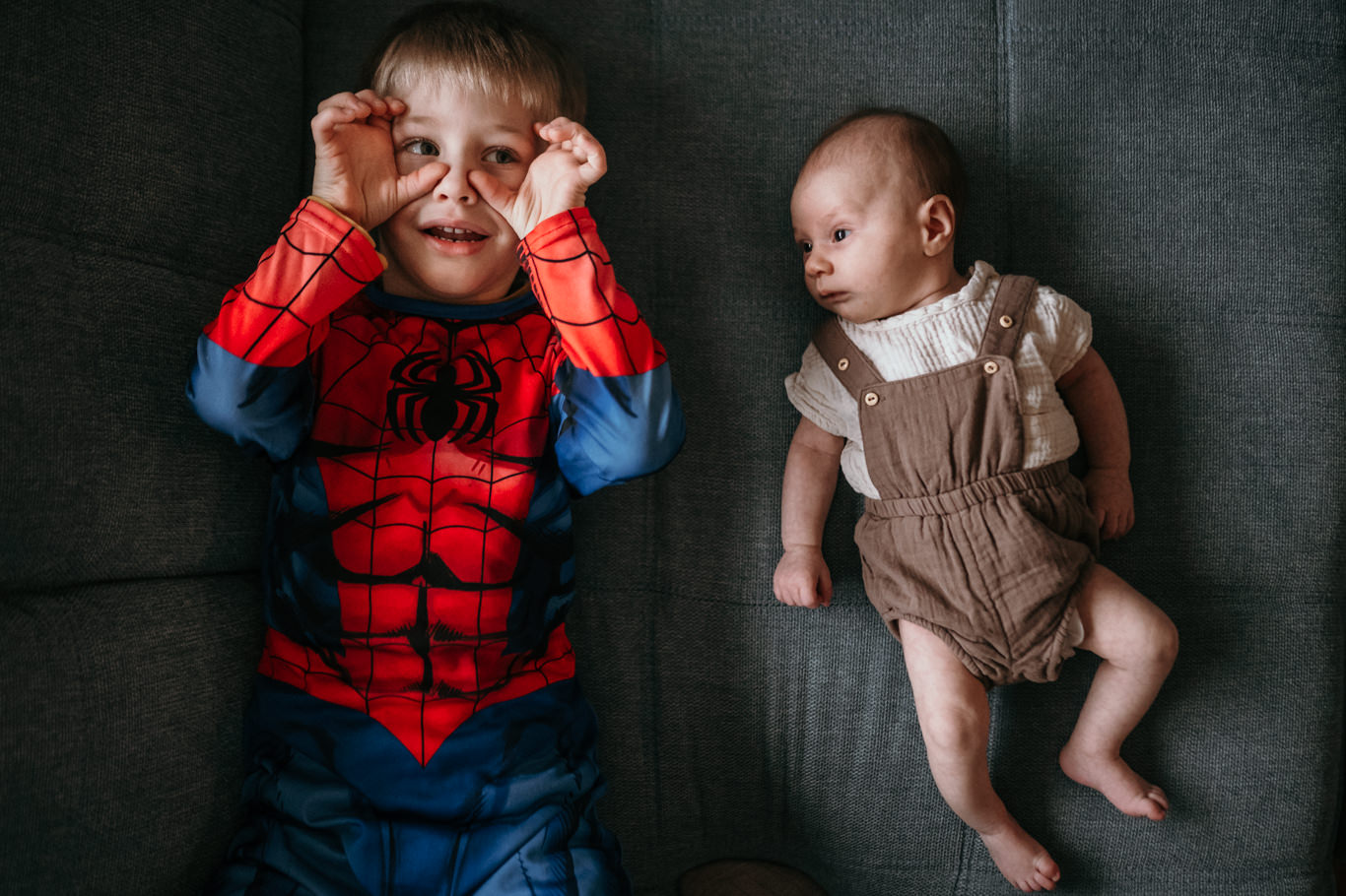 Kleiner Junge im Spiderman-Kostüm liegt neben seinem neugeborenen Geschwisterchen