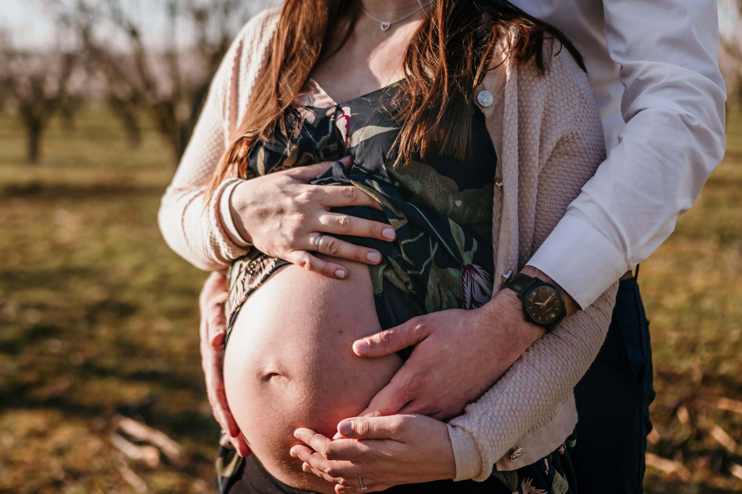 Schwangere hält ihren nackten Bauch, im Hintergrund ein Obstgarten
