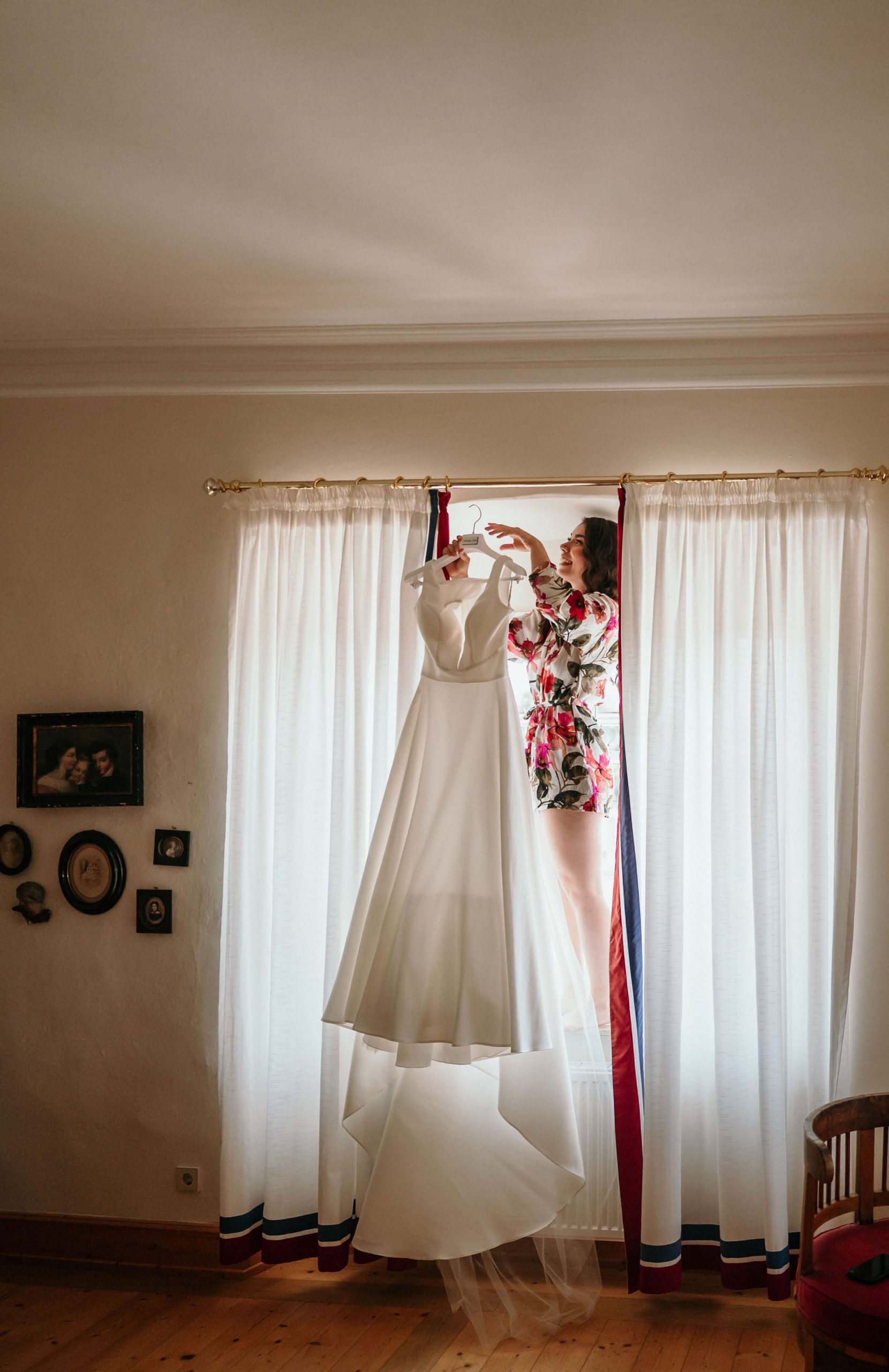 Braut holt Brautkleid von einer Vorhangstange herunter beim Getting Ready während des Hochzeitstags in Schloss Neuhaus in Sinsheim