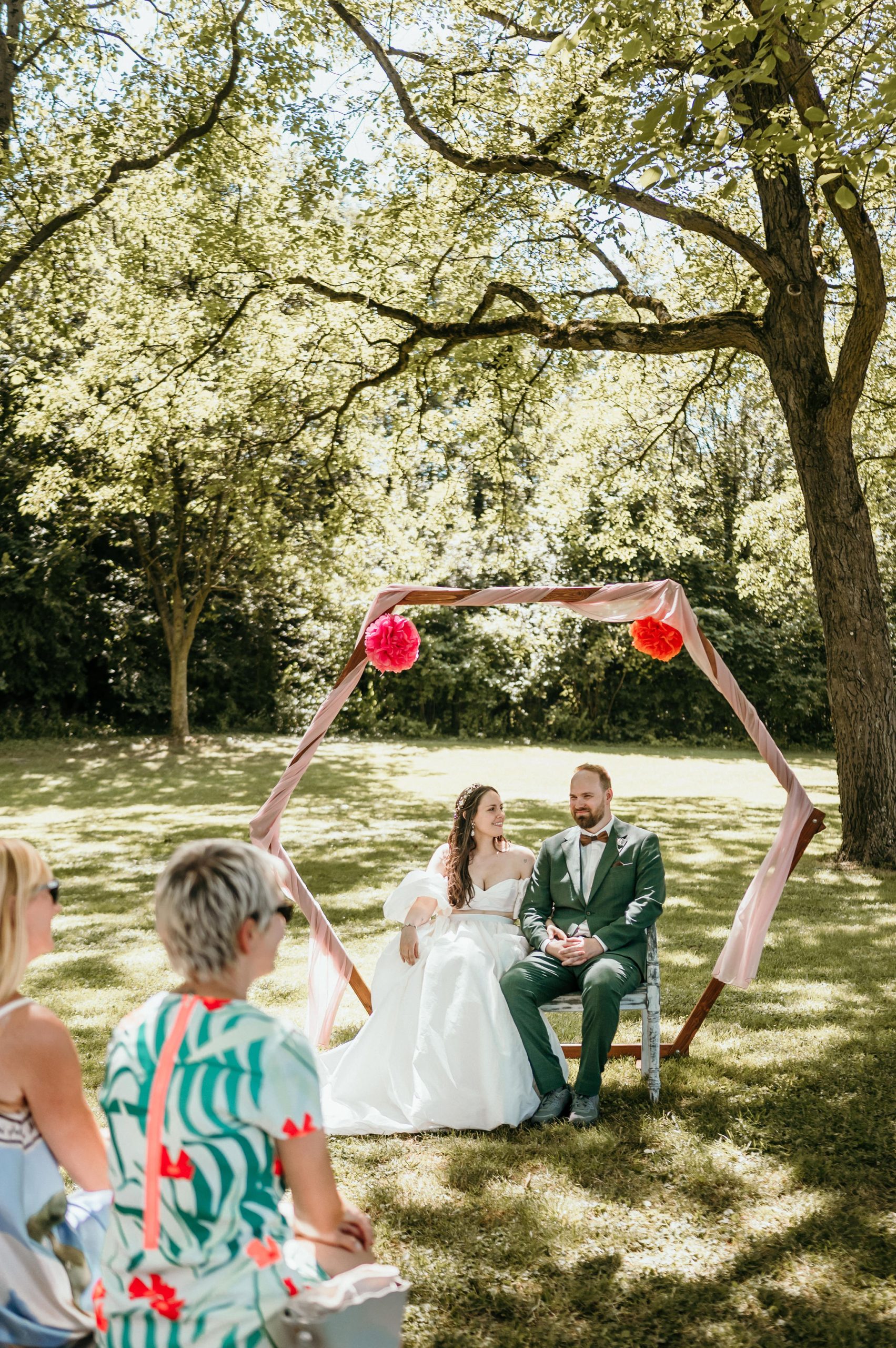 Brautpaar sitzt unter einem Blumenbogen während der Trauung im Freien