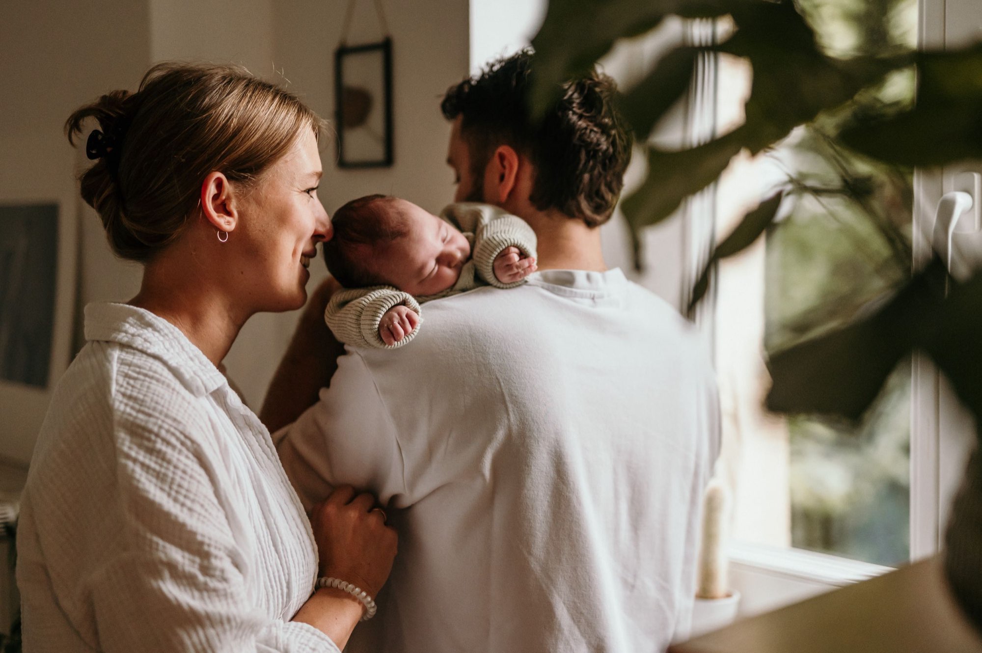 Mutter hält ihr Neugeborenes, Vater steht daneben am Fenster